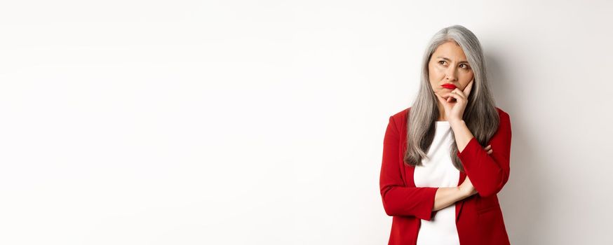 Troubled asian businesswoman in red blazer and lips, pouting and looking left annoyed, standing against white background.