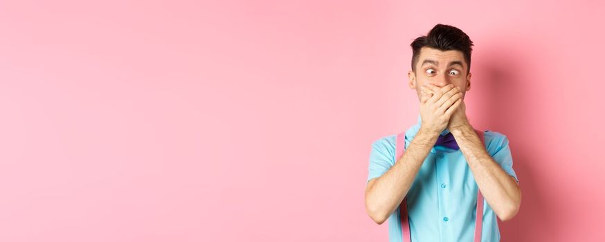 Funny young man covering mouth with hands and squint eyes, making clown grimaces, standing silly on pink background, entertain people.