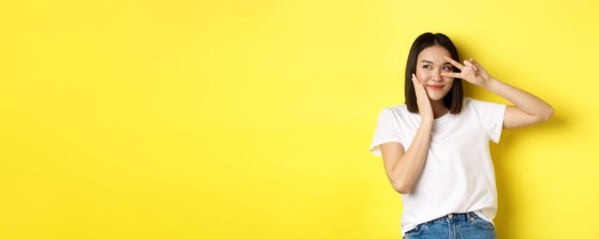 Lovely asian girl in white t-shirt posing with hand on cheek, showing peace sign on eye, standing over yellow background.