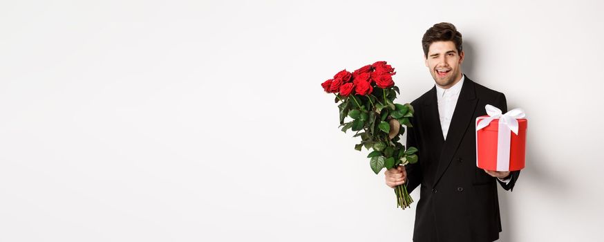 Concept of holidays, relationship and celebration. Handsome and confident man in black suit, going on a date, holding bouquet of roses and present, standing against white background.