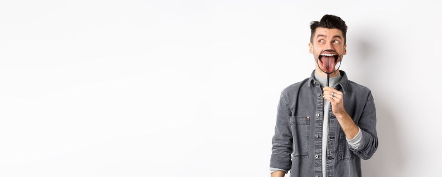 Handsome positive guy showing white perfect teeth and tongue with magnifying glass, looking left at logo, standing against white background.