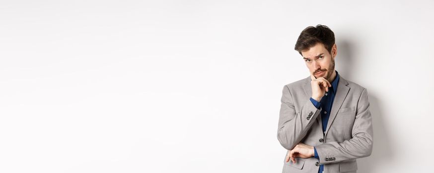 Hesitant businessman in suit making decision, looking pensive at camera with concerned face, having doubts, standing on white background.