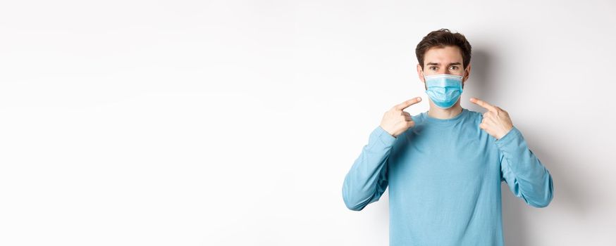 Coronavirus, health and quarantine concept. Young caucasian man in casual clothes pointing at his medical mask, asking to use preventive measures during pandemic, white background.