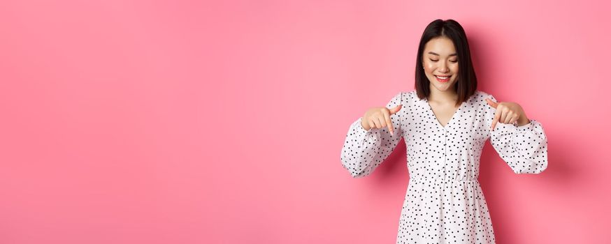 Cute romantic asian woman smiling, pointing fingers and looking down with pleased face, showing product discount, standing over pink background.