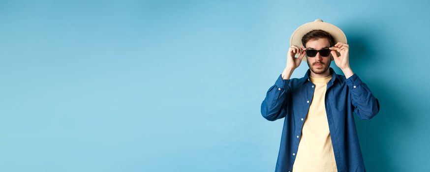 Handsome tourist in straw hat put on sunglasses on summer vacation, standing on blue background.