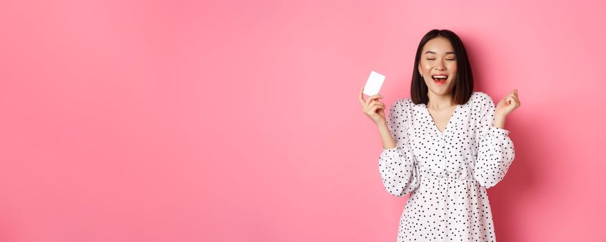 Shopping concept. Excited asian woman feeling satisfaction, holding credit card and rejoicing, standing over pink background.