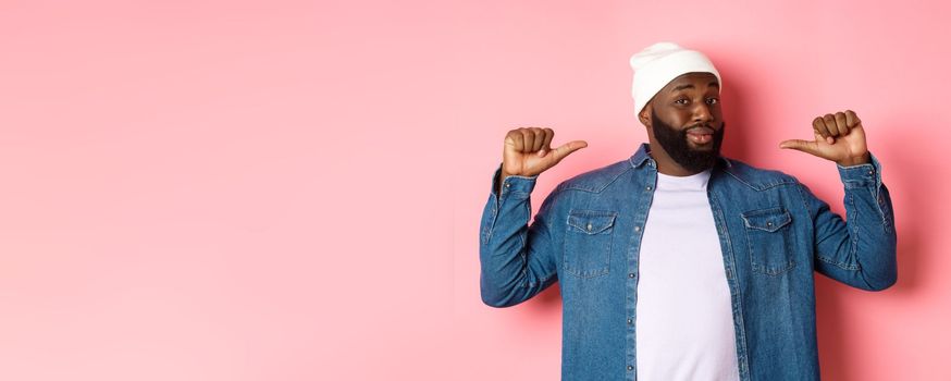 Confident and cheeky Black man pointing at himself, staring at camera self-assured, show-off, standing over pink background.