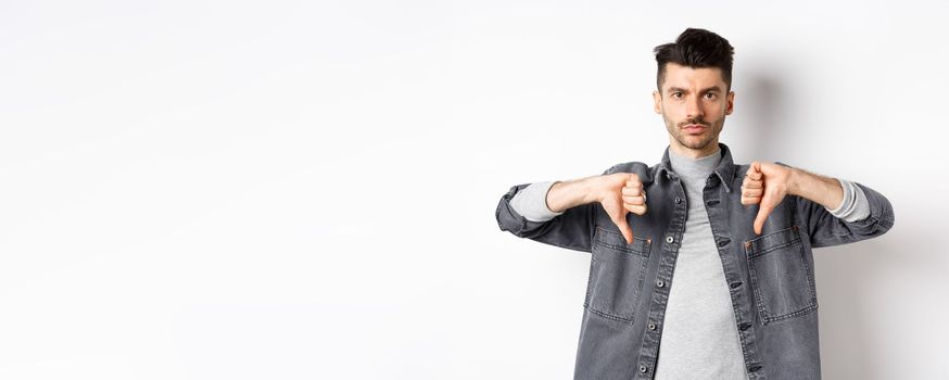 Very bad. Serious man showing thumbs down and look at camera with pokerface, telling no, disapprove and dislike product, standing dissatisfied on white background.