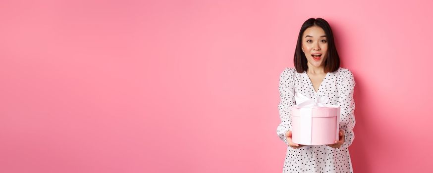 Cute asian woman congratulate with holiday or birthday, giving gift in cute box, standing over pink background.