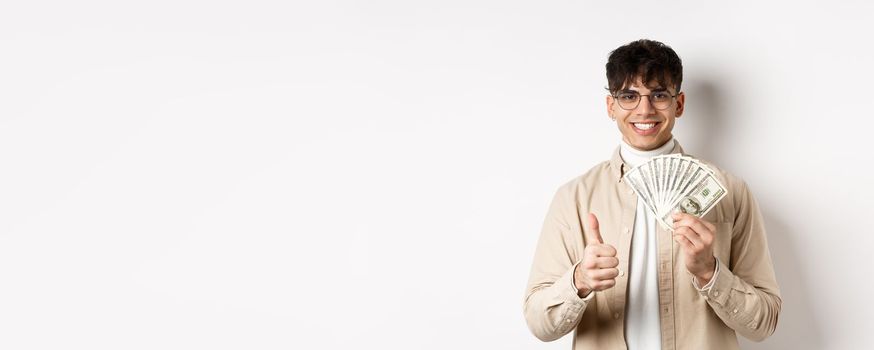 Young guy in glasses showing dollar bills and thumbs up, making money, standing with cash on white background.