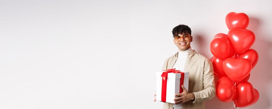 Valentines day and romance concept. Man in love prepare surprise gift for lover, holding present in box and standing near red hearts gesture, white background.