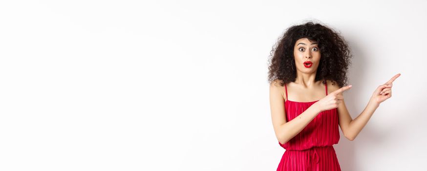 Surprised woman in red dress and makeup pointing fingers right, showing logo and look intrigued, standing on white background.