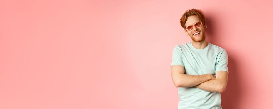 Tourism and vacation concept. Friendly young man with red hair, wearing sunglasses and t-shirt, cross arms on chest and smiling joyfully at camera, pink background.