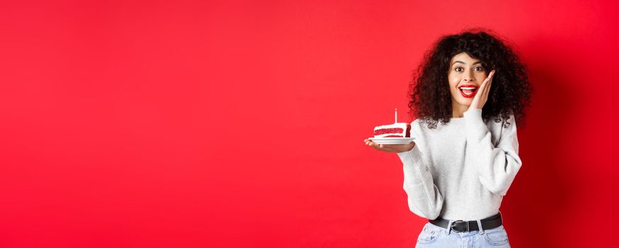 Excited woman celebrating birthday, holding cake and looking surprised and happy at camera, red background.