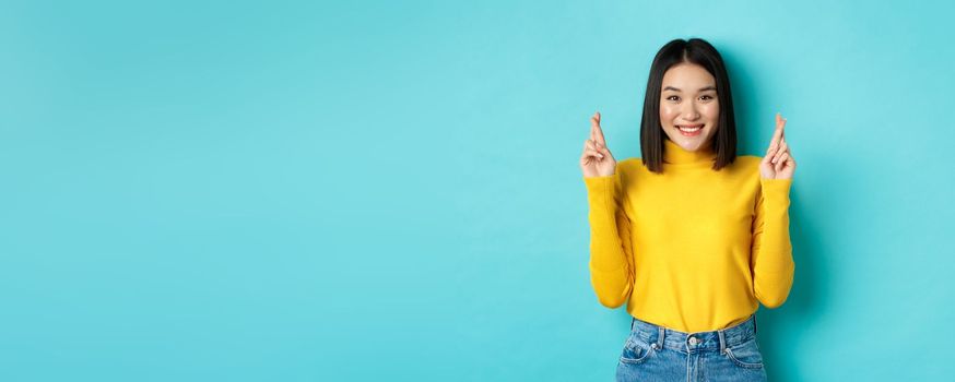 Hopeful and optimistic asian woman cross fingers for good luck, making wish and smiling, looking at camera and expecting something, standing over blue background.