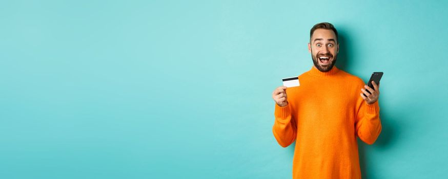 Online shopping. Surprised man holding mobile phone and credit card, paying in internet store, standing over light blue background.