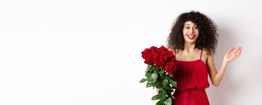 Romance and Valentines day. Woman gasping surprised and happy, receive surprise gift from lover, holding bouquet of red roses, standing on white background.