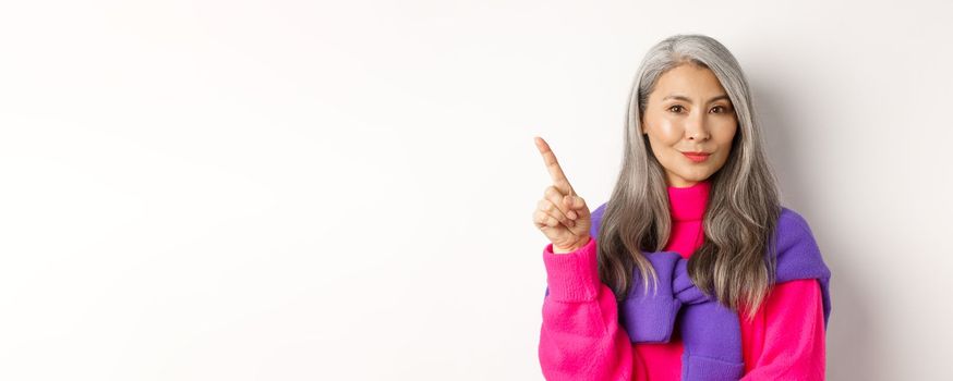 Close-up of asian beautiful mother with grey hair, smiling and pointing upper left corner, showing advertisement, standing over white background.
