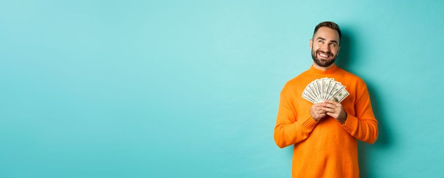 Man thinking about shopping, holding money and smiling satisfied, looking at upper left corner, standing over light blue background.