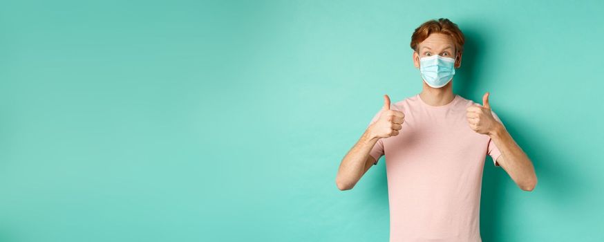 Covid-19, pandemic and lifestyle concept. Cheerful redhead guy in medical mask showing thumbs up in approval, like and praise product, standing over turquoise background.