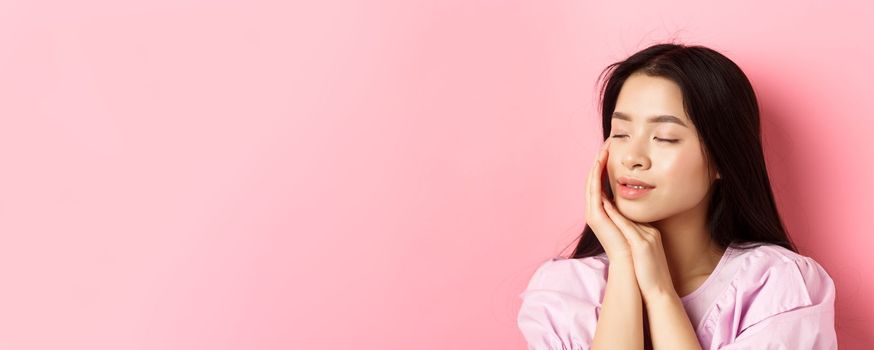 Tender and romantic girl gently touching face, close eyes and smiling softly, standing on pink background. Concept of skincare.