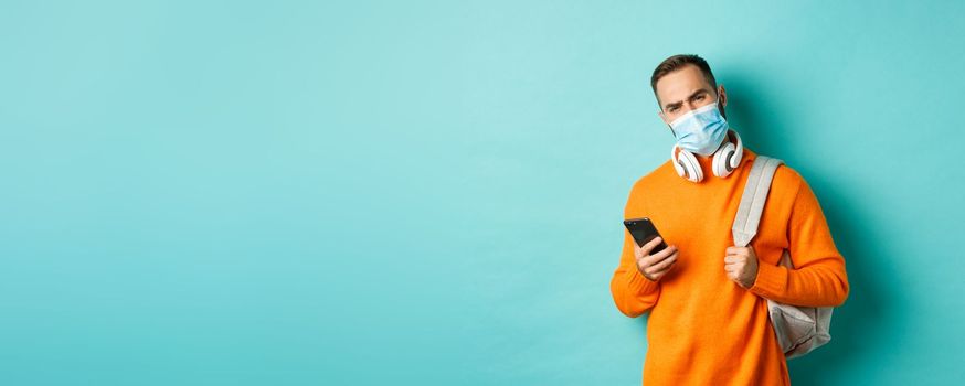 Skeptical and disappointed young man wearing face mask, holding backpack and mobile phone, frowning upset, standing over light blue background.