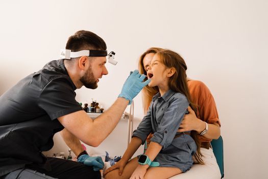 Oropharyngoscopy family procedure for child. Mother with daughter visit ENT doctor. Otolaryngologist examines child throat with spatula. Family consultation with laryngologist