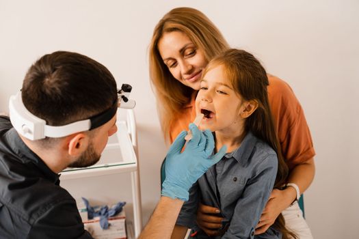 Oropharyngoscopy family procedure for child. Mother with daughter visit ENT doctor. Otolaryngologist examines child throat with spatula. Family consultation with laryngologist