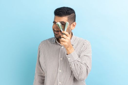 Portrait of curious positive funny businessman peeping through bunch of euro banknotes, passive income, wearing striped shirt. Indoor studio shot isolated on blue background.