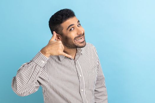 You contact us. Portrait of positive businessman with beard making gesture with fingers dial my number or call me back, wearing striped shirt. Indoor studio shot isolated on blue background.