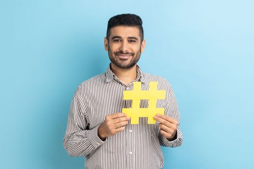 Viral hashtag and successful blogging. Satisfied businessman with beard holding large yellow hash symbol, wearing striped shirt. Indoor studio shot isolated on blue background.