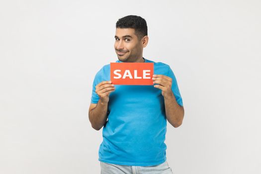 Portrait of positive unshaven man wearing blue T- shirt standing holding sale card in hands looking at camera with toothy smile, black friday. Indoor studio shot isolated on gray background.
