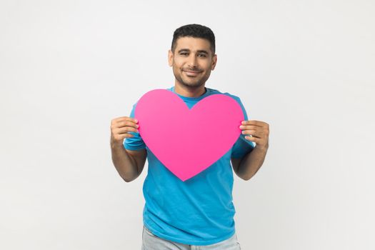 Portrait of romantic smiling joyful unshaven man wearing blue T- shirt standing holding big pink heart, expressing fondness and love. Indoor studio shot isolated on gray background.