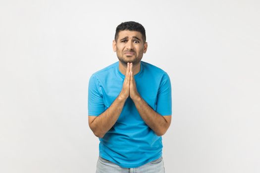 Portrait of sad upset unshaven man wearing blue T- shirt standing has sorrowful expression asking somebody to forgive, keeps palms in praying gesture. Indoor studio shot isolated on gray background.