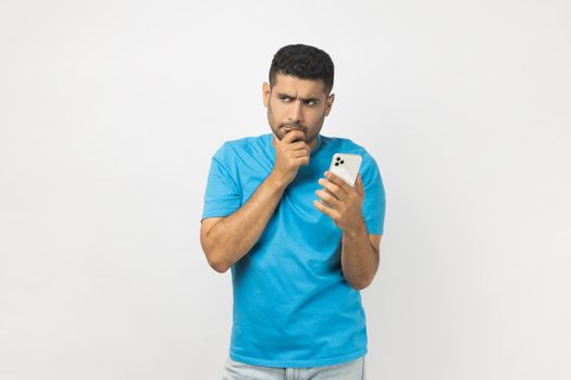 Portrait of pensive thoughtful unshaven man blogger wearing blue T- shirt standing using cell phone, writing post, thinking about answer the message. Indoor studio shot isolated on gray background.