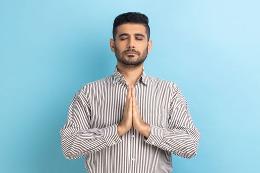 Portrait of calm relaxed young businessman with beard standing doing yoga meditating exercise, keeping palms together, wearing striped shirt. Indoor studio shot isolated on blue background.