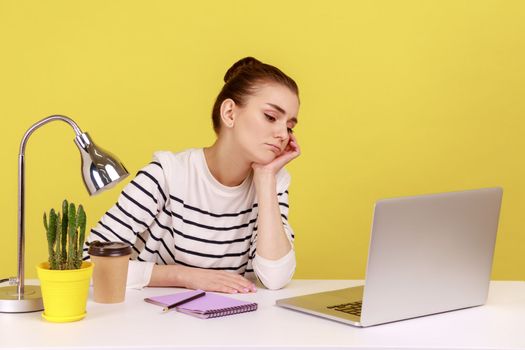 Bored tired woman manager sitting at workplace and looking at laptop screen, leaning on hand, feeling lazy uninterested in her job. Indoor studio studio shot isolated on yellow background.