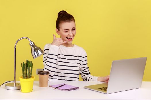Call me. Beautiful lovely young woman employee sitting at workplace with laptop keeping hand shaped like telephone near head. Indoor studio studio shot isolated on yellow background.