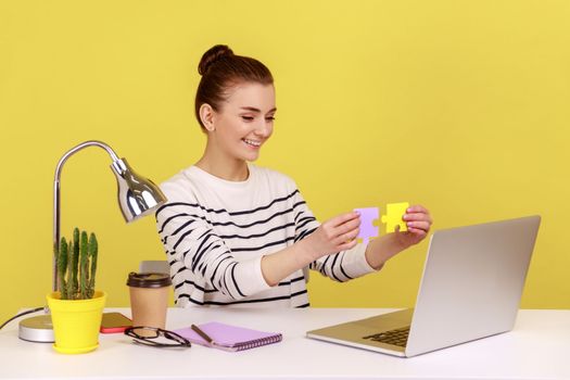 Satisfied woman showing to laptop display two colored pieces of jigsaw, easily solving difficult tasks at work, sitting at workplace. Indoor studio studio shot isolated on yellow background.