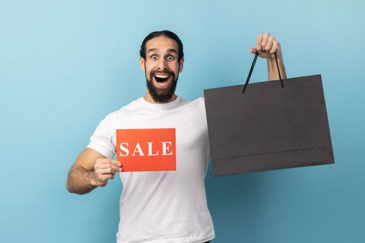 Portrait of shocked man with beard wearing white T-shirt holding shopping bags and showing sale card, advertising low prices in the mall. Indoor studio shot isolated on blue background.