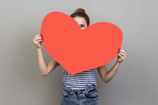 Portrait of woman hiding face behind big red heart and looking at camera with curious prying eyes, expressing i love you, feeling affection fondness. Indoor studio shot isolated on gray background.