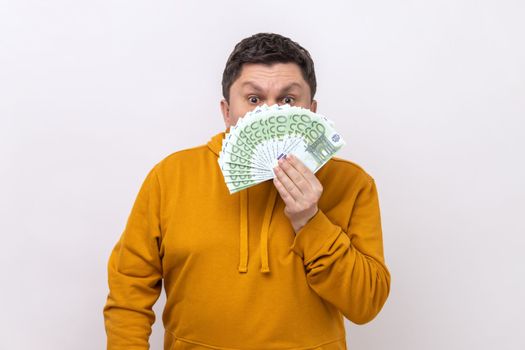 Dark haired man hiding half of face behind fan of hundred euro bills, big winning, betting, wearing urban style hoodie. Indoor studio shot isolated on white background.