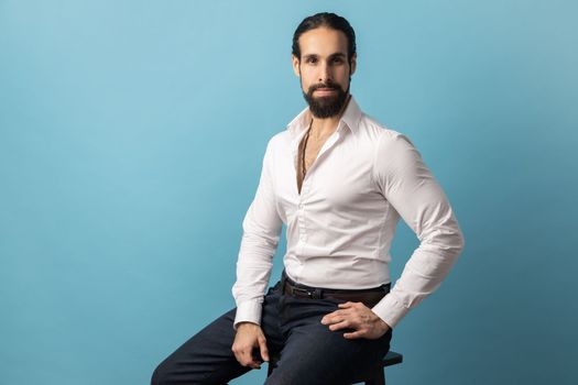 Portrait of serious handsome man with beard and collected dark wearing white shirt looking at camera with confident facial expression. Indoor studio shot isolated on blue background.