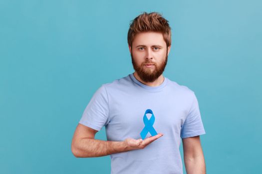 Portrait of serious young adult attractive bearded man holding blue awareness, disease symbol, looking at camera, support and care. Indoor studio shot isolated on blue background.
