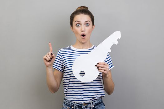 Portrait of surprised woman wearing striped T-shirt holding big white house key, real estate advertisement, pointing finger up, having excellent idea. Indoor studio shot isolated on gray background.