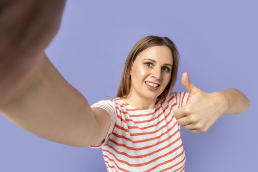 Portrait of delighted beautiful blond woman wearing striped T-shirt taking selfie photo, holding smartphone camera and showing thumb up, POV. Indoor studio shot isolated on purple background.