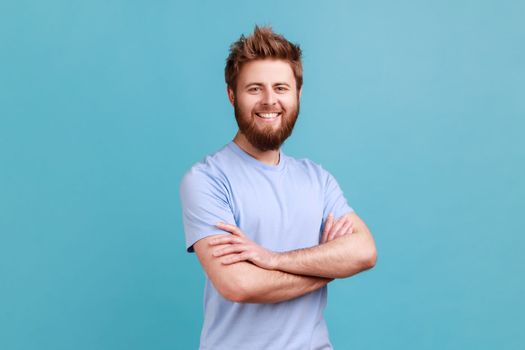 Portrait of satisfied happy bearded man standing with crossed arms and looking at camera with toothy smile, having positive confident expression. Indoor studio shot isolated on blue background.