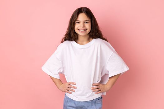 Portrait of delighted smiling little girl wearing white T-shirt standing hands on hips, looking at camera with happy expression. Indoor studio shot isolated on pink background.