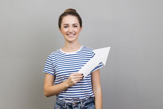 Portrait of satisfied delighted smiling woman in striped T-shirt showing white paper arrow indicating aside, looking at camera with positive emotions. Indoor studio shot isolated on gray background.
