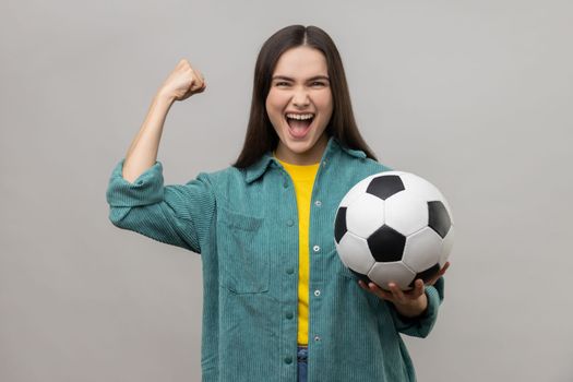Satisfied woman screaming widely opening mouth, celebrating victory of favourite football team, holding ball in hands, wearing casual style jacket. Indoor studio shot isolated on gray background.